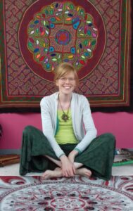 Who is Art Sound & Medicine Woman? Frouke Vermeulen sitting cross-legged and smiling in front of a large Shipibo altar fabric hanging on the wall.