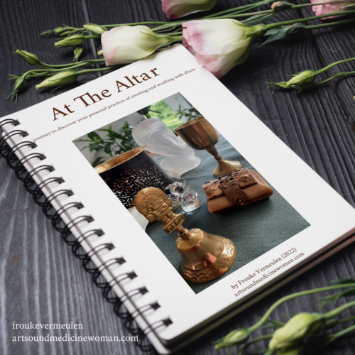A spiral bound A4 notebook surrounded by white roses with a pink touch. On the cover of the notebook is a picture of an altar holding sacred objects such as a selenite cobra, a bell from the Queros in Peru, a mini treasure box etc.
