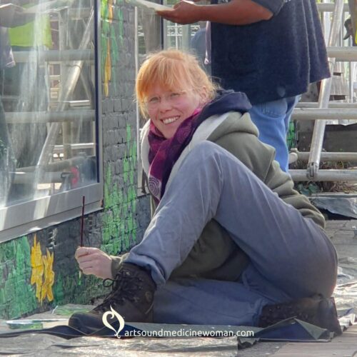 Frouke Vermeulen smiling at the camera, sitting down in a funny position while painting a mural at Groenplaats in Zoerle-Parwijs, Belgium.