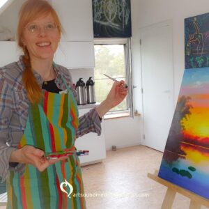 Frouke Vermeulen smiling at the camera, holding a brush in her left hand, a palette in her right hand, while standing in front of her painting.